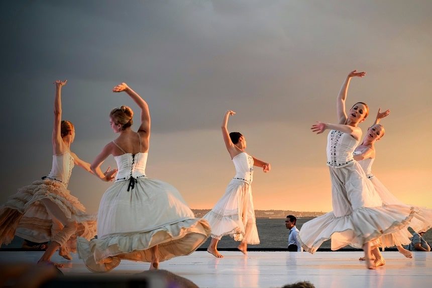 Five young women dancing in a circle. All are looking to their left with their right arm raised and the left arm curved in from of them.