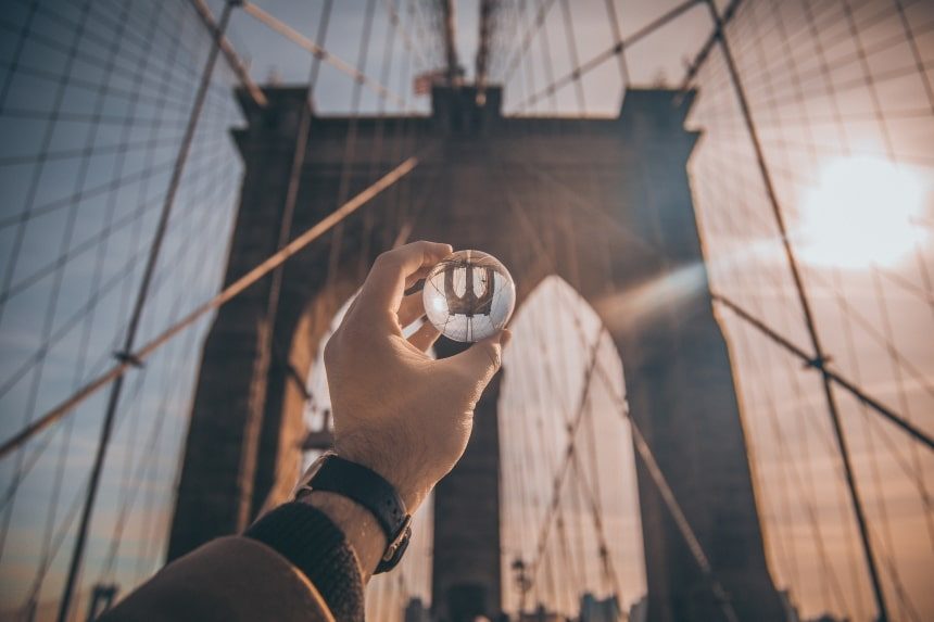 A male hand is holding a glass ball up to the Brooklyn Bridge in New York. The bridge is reflected upside down in the glass ball.