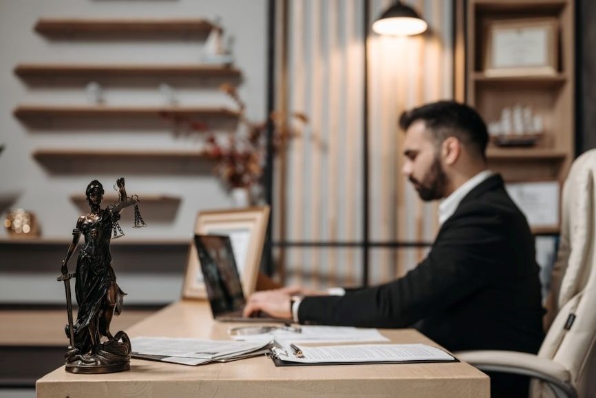 A man in black suit sitting at a desk using a laptop. With a lady justice statue on the end of the desk. 