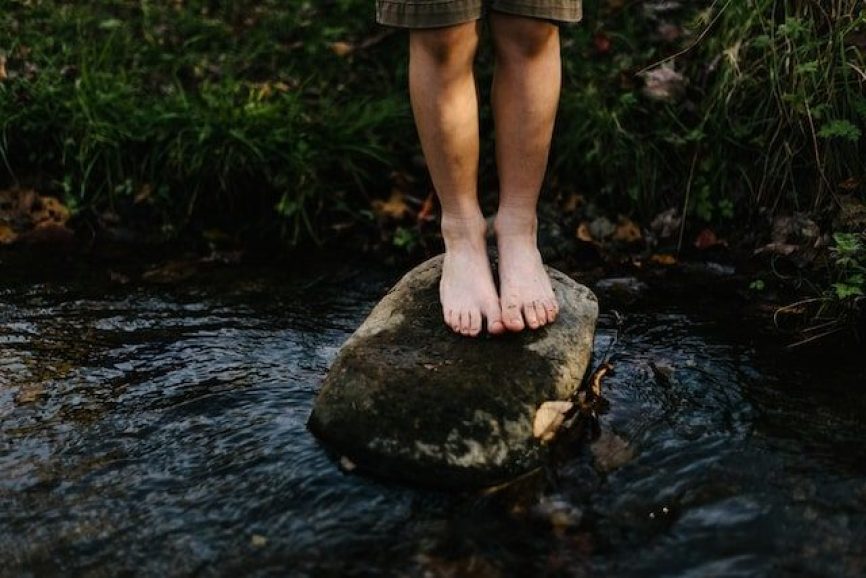 Close up of a person’s feet standing on a rock in a stream.