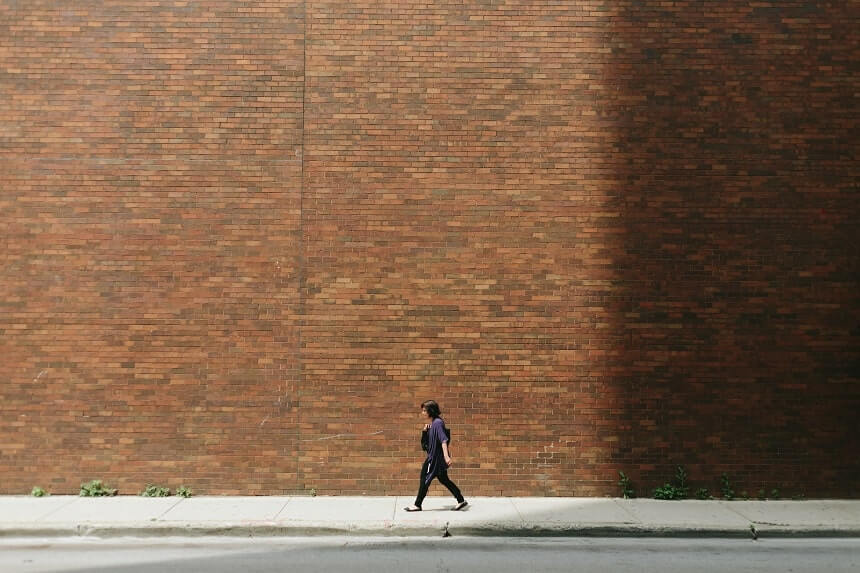 Image, dark haired human walking past a very high brick wall