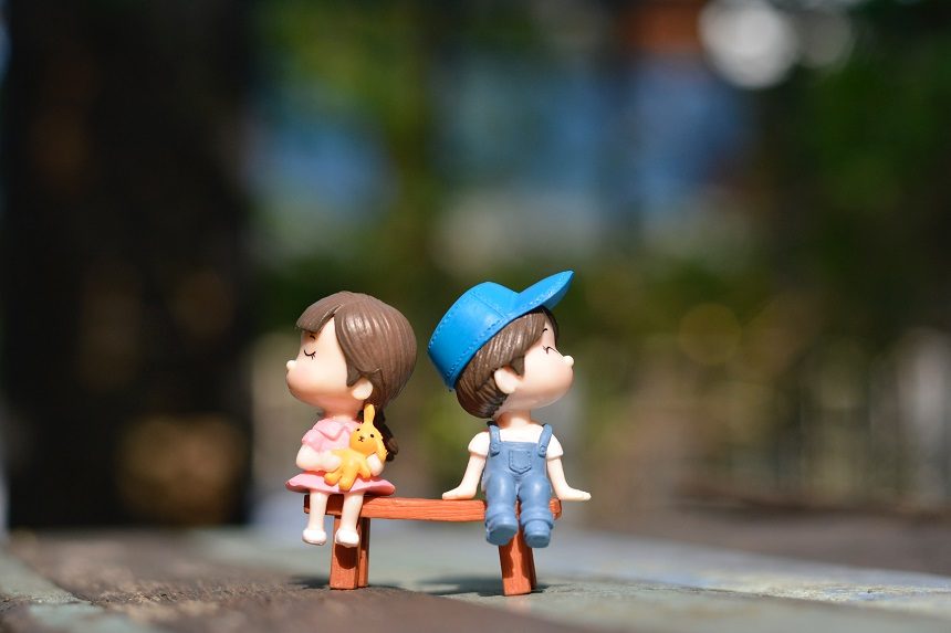 Image, a toy girl and boy sitting on a small table facing away from each other looking grumpy.