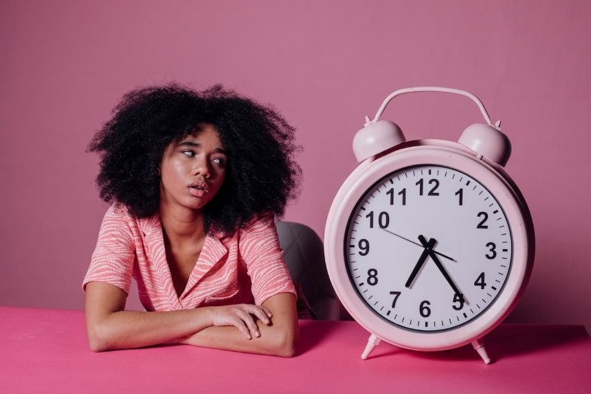 Woman sitting next to and looking at a large pink clock with a worried expression. 
