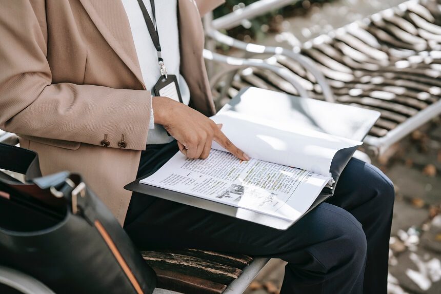 A person dressed in business clothes sits on a bench, their finger pointing to text in the bound article on their lap