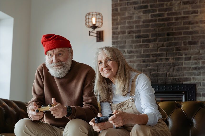 A man with a grey beard and a woman with grey hair both using Playstation controllers and smiling.