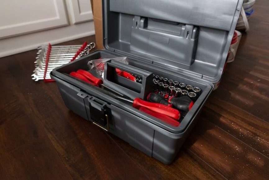 A grey plastic toolbox on a wooden floor, filled with red-handled tools and attachments.