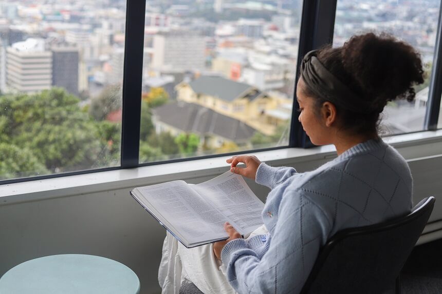Looking over a young woman’s shoulder as she reads an academic journal. She sits next to a window in a high-rise building looking over the city