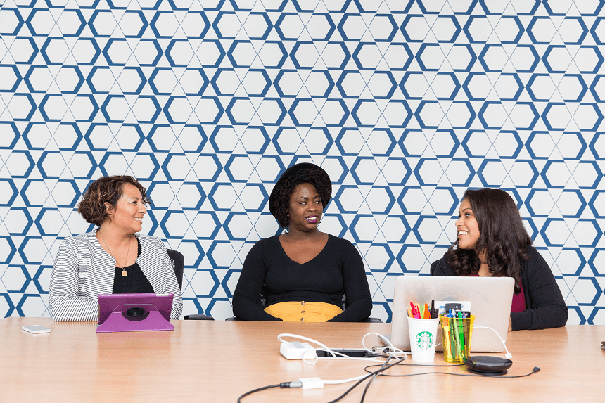 Three femme-presenting people sitting at a table with laptops and other office-type things.